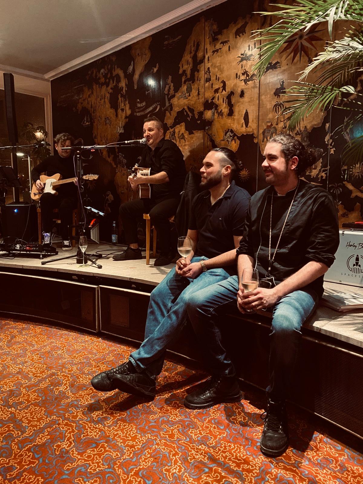 Andy Gas und sein Partner sitzen am Bühnenrand, während hinter ihnen eine Band live spielt